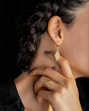 A close-up of a person's neck, ear, and hand. The person is wearing gold jewelry, including an intricate earring and matching ring, both with a similar geometric, bead-like design. The hair is styled in braids, and the person is wearing dark nail polish.