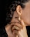 A close-up of a person's neck, ear, and hand. The person is wearing gold jewelry, including an intricate earring and matching ring, both with a similar geometric, bead-like design. The hair is styled in braids, and the person is wearing dark nail polish.