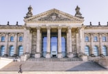 A large neoclassical building with prominent columns and a pediment adorned with elaborate sculptures. The facade features the inscription 'DEM DEUTSCHEN VOLKE' above the entrance. Wide stone steps lead up to the entrance, with people visible walking and sitting on them.