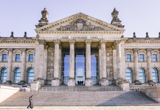 A large neoclassical building with prominent columns and a pediment adorned with elaborate sculptures. The facade features the inscription 'DEM DEUTSCHEN VOLKE' above the entrance. Wide stone steps lead up to the entrance, with people visible walking and sitting on them.