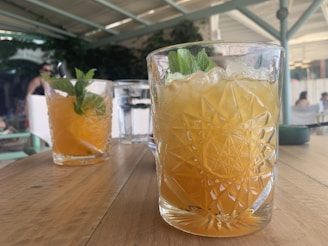 Two glasses of an orange-colored beverage with mint leaves sit on a wooden table. The glasses have a textured, patterned design. In the background, a partially visible glass of water and some people are seated in a shaded, outdoor setting.