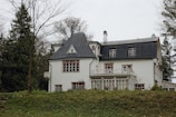 A white, two-story house with a gray, sloped roof is surrounded by trees. The building features multiple windows with brown frames and a wraparound balcony. The yard is grassy with scattered, bare trees, indicating a winter or early spring setting.
