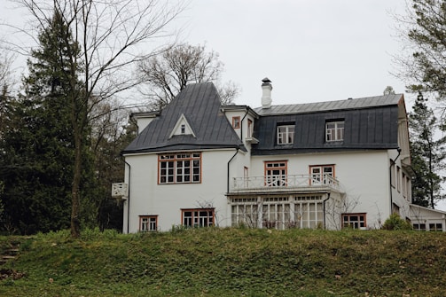 A white, two-story house with a gray, sloped roof is surrounded by trees. The building features multiple windows with brown frames and a wraparound balcony. The yard is grassy with scattered, bare trees, indicating a winter or early spring setting.