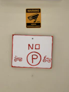 A yellow sign with a black image of a surveillance camera and the words 'WARNING VIDEO SURVEILLANCE 24H' is placed above a red and white sign. The lower sign displays a red 'NO P' with illustrations of motorcycles on each side.