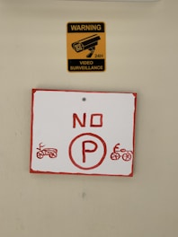A yellow sign with a black image of a surveillance camera and the words 'WARNING VIDEO SURVEILLANCE 24H' is placed above a red and white sign. The lower sign displays a red 'NO P' with illustrations of motorcycles on each side.
