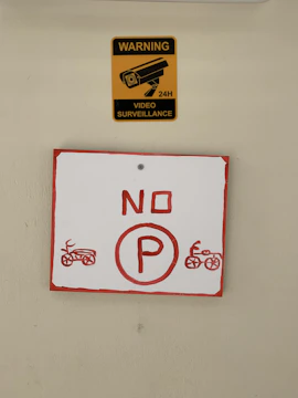 A yellow sign with a black image of a surveillance camera and the words 'WARNING VIDEO SURVEILLANCE 24H' is placed above a red and white sign. The lower sign displays a red 'NO P' with illustrations of motorcycles on each side.