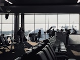 A busy airport gate area features several silhouetted figures, including passengers with luggage and airport staff interacting at a counter. Rows of empty seats fill the foreground and natural light streams through large windows, partially obscured by the visible nose of an airplane.