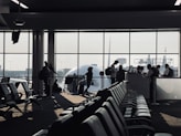 A busy airport gate area features several silhouetted figures, including passengers with luggage and airport staff interacting at a counter. Rows of empty seats fill the foreground and natural light streams through large windows, partially obscured by the visible nose of an airplane.