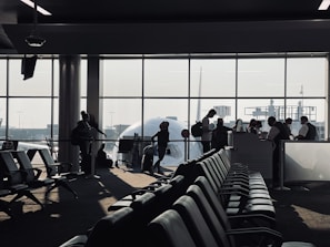 A busy airport gate area features several silhouetted figures, including passengers with luggage and airport staff interacting at a counter. Rows of empty seats fill the foreground and natural light streams through large windows, partially obscured by the visible nose of an airplane.