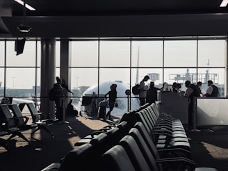 A busy airport gate area features several silhouetted figures, including passengers with luggage and airport staff interacting at a counter. Rows of empty seats fill the foreground and natural light streams through large windows, partially obscured by the visible nose of an airplane.