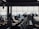 A busy airport gate area features several silhouetted figures, including passengers with luggage and airport staff interacting at a counter. Rows of empty seats fill the foreground and natural light streams through large windows, partially obscured by the visible nose of an airplane.