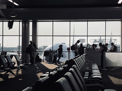 A busy airport gate area features several silhouetted figures, including passengers with luggage and airport staff interacting at a counter. Rows of empty seats fill the foreground and natural light streams through large windows, partially obscured by the visible nose of an airplane.