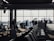 A busy airport gate area features several silhouetted figures, including passengers with luggage and airport staff interacting at a counter. Rows of empty seats fill the foreground and natural light streams through large windows, partially obscured by the visible nose of an airplane.