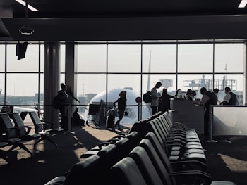 A busy airport gate area features several silhouetted figures, including passengers with luggage and airport staff interacting at a counter. Rows of empty seats fill the foreground and natural light streams through large windows, partially obscured by the visible nose of an airplane.
