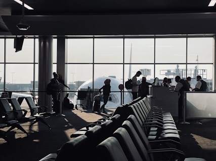 A busy airport gate area features several silhouetted figures, including passengers with luggage and airport staff interacting at a counter. Rows of empty seats fill the foreground and natural light streams through large windows, partially obscured by the visible nose of an airplane.