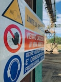 A close-up of a safety sign with warnings and instructions, including 'Limite di sicurezza' and 'Vietato l'accesso ai non addetti'. The sign features symbols such as a hand indicating 'no access' and an icon of headphones with a message about hearing protection. The background shows an outdoor industrial setting.