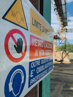 A close-up of a safety sign with warnings and instructions, including 'Limite di sicurezza' and 'Vietato l'accesso ai non addetti'. The sign features symbols such as a hand indicating 'no access' and an icon of headphones with a message about hearing protection. The background shows an outdoor industrial setting.