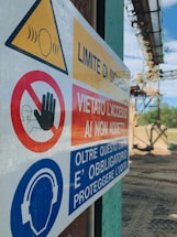 A close-up of a safety sign with warnings and instructions, including 'Limite di sicurezza' and 'Vietato l'accesso ai non addetti'. The sign features symbols such as a hand indicating 'no access' and an icon of headphones with a message about hearing protection. The background shows an outdoor industrial setting.