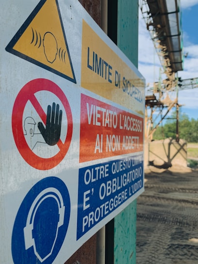A close-up of a safety sign with warnings and instructions, including 'Limite di sicurezza' and 'Vietato l'accesso ai non addetti'. The sign features symbols such as a hand indicating 'no access' and an icon of headphones with a message about hearing protection. The background shows an outdoor industrial setting.