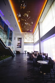 A modern, spacious lobby area with high ceilings and large windows allowing natural light to fill the room. Geometric light fixtures and wooden panels on the ceiling add a stylish touch. A staircase with glass railings leads to an upper level. Several people are seated at tables, engaged in conversation.