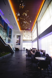 A modern, spacious lobby area with high ceilings and large windows allowing natural light to fill the room. Geometric light fixtures and wooden panels on the ceiling add a stylish touch. A staircase with glass railings leads to an upper level. Several people are seated at tables, engaged in conversation.