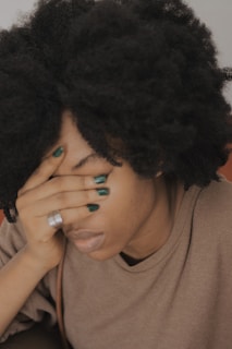A person with their hand covering their face, possibly expressing distress or fatigue. The individual has curly hair and is wearing a simple, comfortable outfit. The lighting is soft, creating a calm atmosphere.