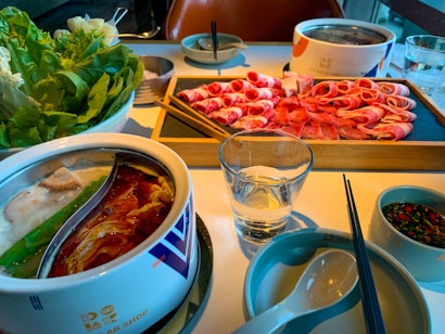 A table set for a hot pot meal with a dual-section pot containing broth, surrounded by dishes of fresh green vegetables, thinly sliced raw meat, dipping sauces, and utensils. The dining setup suggests a shared meal experience.