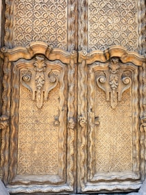 Intricately carved wooden doors feature elaborate geometric patterns and ornate floral motifs. The wood appears aged, showcasing natural textures and a warm color, enhancing the craftsmanship and artistic detailing on the surface.