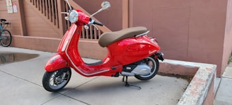 A bright red scooter with a brown seat is parked on a concrete surface near a peach-colored wall. The background includes stairs and another parked bicycle. The scooter is glossy with chrome accents and rests on its kickstand.