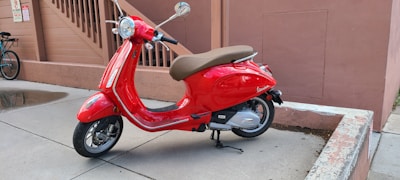 A bright red scooter with a brown seat is parked on a concrete surface near a peach-colored wall. The background includes stairs and another parked bicycle. The scooter is glossy with chrome accents and rests on its kickstand.
