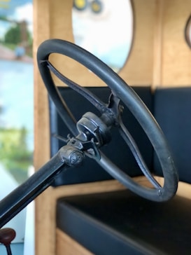 A vintage car steering wheel is prominently positioned with a simple, classic design. The background shows blurred elements with light wooden tones and some green and blue hues, suggesting an indoor setting or a vintage car exhibition.