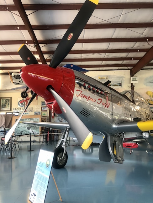 A vintage aircraft with a polished metallic body and red accents is displayed in an indoor setting. The propeller has three black blades with yellow tips. On the fuselage, the words 'Tempus Fugit' are written. There are various aviation-related posters, paintings, and a signboard near the aircraft, indicating it might be an aviation museum.