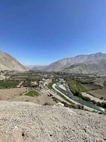 A vast landscape features a river winding through a valley surrounded by mountainous terrain. The river is flanked by lush green fields and sparse settlements. The sky is clear and blue, creating a contrast with the earthy tones of the mountains and fields.