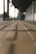 A blurred perspective of a walkway with tactile paving in the foreground and a covered walkway structure in the background. The scene appears tranquil and spacious, with subtle lighting suggesting an early morning or late afternoon setting.