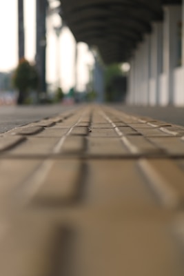 A blurred perspective of a walkway with tactile paving in the foreground and a covered walkway structure in the background. The scene appears tranquil and spacious, with subtle lighting suggesting an early morning or late afternoon setting.