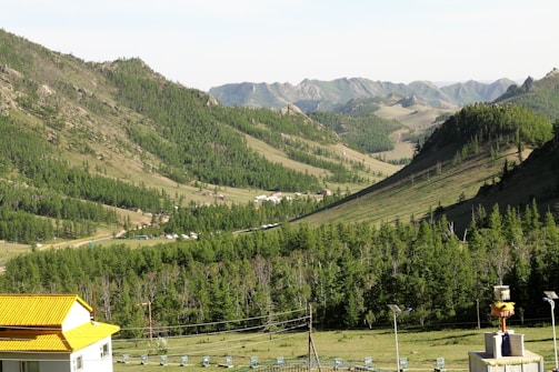 A lush green valley surrounded by rolling hills and mountains is visible. The area is dotted with patches of dense forest and scattered small buildings. A modern, white house with a bright yellow roof stands in the foreground. The landscape stretches into the distance, with the blue sky meeting the distant hills.
