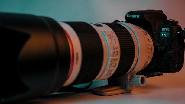 A professional Canon camera with a large telephoto lens is placed on a surface. The lighting is a mix of teal and warm tones, highlighting the lens and camera body. The surface appears smooth and the background is softly blurred, adding focus to the camera setup.