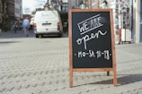A wooden-framed chalkboard with handwritten message 'We are open MO-SA 10-18' is placed on a cobblestone street. In the background, a parked van and pedestrians walking along the sidewalk suggest an urban setting.