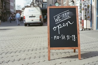 A wooden-framed chalkboard with handwritten message 'We are open MO-SA 10-18' is placed on a cobblestone street. In the background, a parked van and pedestrians walking along the sidewalk suggest an urban setting.