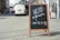 A wooden-framed chalkboard with handwritten message 'We are open MO-SA 10-18' is placed on a cobblestone street. In the background, a parked van and pedestrians walking along the sidewalk suggest an urban setting.