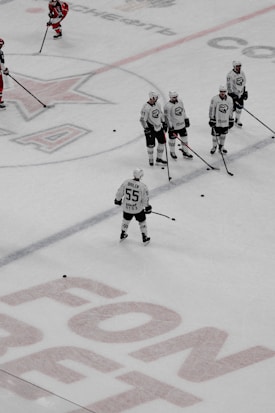 A group of ice hockey players in white uniforms are standing on an ice rink, possibly during a practice or warm-up session. One player, separated from the group, is facing away from the camera. Several pucks are scattered on the ice, and a large logo and text are visible on the rink surface. In the background, a few players in red uniforms can be seen.