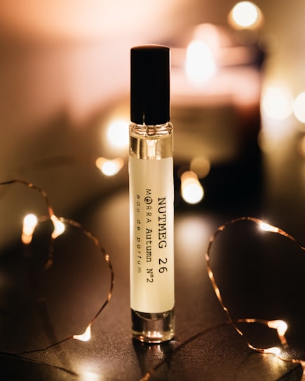 A perfume bottle labeled 'Nutmeg 26 MORRA Autumn No.2 eau de parfum' stands upright on a dark surface. Soft, warm lighting surrounds the bottle, creating a cozy and intimate atmosphere. Small, fairy lights are elegantly arranged around the bottle, adding a touch of sparkle and enhancing the ambient glow.