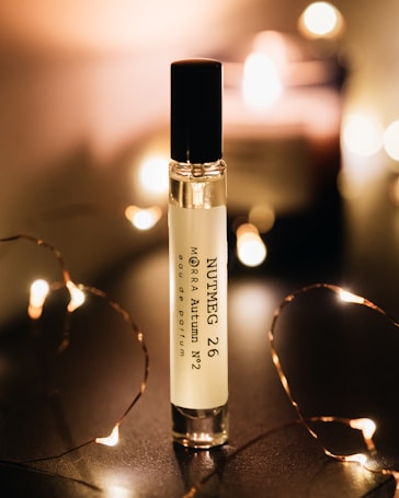 A perfume bottle labeled 'Nutmeg 26 MORRA Autumn No.2 eau de parfum' stands upright on a dark surface. Soft, warm lighting surrounds the bottle, creating a cozy and intimate atmosphere. Small, fairy lights are elegantly arranged around the bottle, adding a touch of sparkle and enhancing the ambient glow.