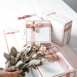 A collection of elegantly wrapped gift boxes in white with pink ribbons, arranged on a white surface. In the foreground, a hand is holding a bouquet of pink roses, complementing the color scheme of the gift boxes.
