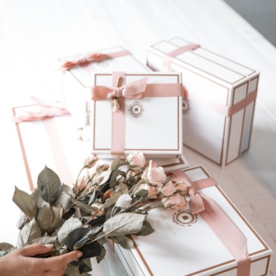 A collection of elegantly wrapped gift boxes in white with pink ribbons, arranged on a white surface. In the foreground, a hand is holding a bouquet of pink roses, complementing the color scheme of the gift boxes.