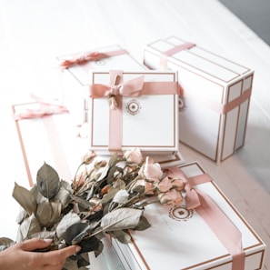 A collection of elegantly wrapped gift boxes in white with pink ribbons, arranged on a white surface. In the foreground, a hand is holding a bouquet of pink roses, complementing the color scheme of the gift boxes.
