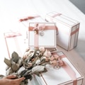 A collection of elegantly wrapped gift boxes in white with pink ribbons, arranged on a white surface. In the foreground, a hand is holding a bouquet of pink roses, complementing the color scheme of the gift boxes.