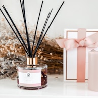 A decorative setup featuring a glass aroma diffuser with black reeds, positioned on a white surface. Next to it is a white gift box with a pink ribbon and a cylindrical pink candle. The background includes dried flowers, adding a rustic and elegant touch.