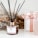 A decorative setup featuring a glass aroma diffuser with black reeds, positioned on a white surface. Next to it is a white gift box with a pink ribbon and a cylindrical pink candle. The background includes dried flowers, adding a rustic and elegant touch.