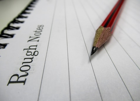 A wooden pencil with a red and black design lies on a lined notebook page. The text 'Rough Notes' is printed on the left side of the notebook. The image conveys a sense of simplicity and focus on writing or note-taking.